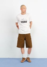 Hvit bomulls t-skjorte med svart "PUMA" grafikk, brune cargo-shorts med lommer, hvite sokker og brune sneakers mot en ensfarget bakgrunn.
