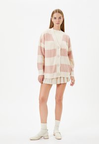 Stickad kofta i krämfärgat och ljust rosa randigt mönster, med V-ringning, knäppning fram och oversize ärmar, stylad med en plisserad kjol.