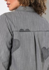 Camicia in denim grigio con una piega sul retro e sottili motivi a forma di cuore in grigio scuro. Il tessuto appare morbido con una texture liscia.