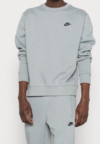 Ljusgrå sweatshirt gjord av mjukt material, med rund hals, ribbade muddar och en liten svart Nike-logotyp på bröstet.