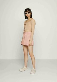 Jovem mulher a usar um crop top de mangas longas bege com atilhos, saia rosa, ténis brancos, óculos de sol pretos, de pé contra um fundo claro liso.