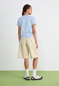 Ljusblå bomulls t-shirt, avslappnad passform. Beiga denimshorts med bakfickor. Krämfärgade strumpor och sneakers med svarta sulor. Grön bakgrund.