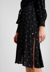 Svart klänning i lätt fabric med blommönster, lång ärm och en sidoöppning, med ett matchande bält vid midjan.