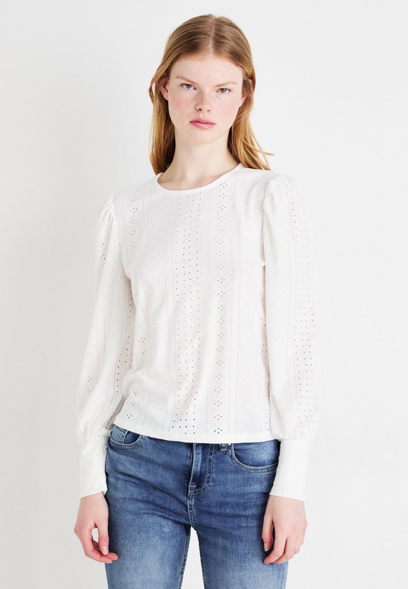 White pullover top with puffed sleeves, featuring embroidered eyelet patterns and a round neckline. Paired with blue denim jeans.