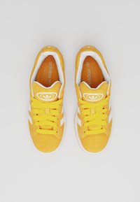 Yellow suede sneakers with white side stripes, round logo on tongue, and textured laces. Features a cushioned white interior.