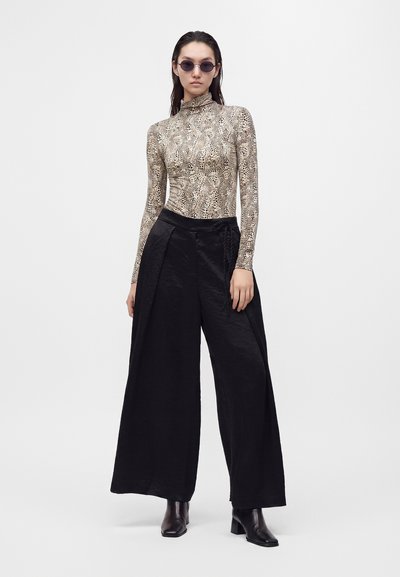 Femme portant des lunettes de soleil rondes foncées, un haut à manches longues col montant avec imprimé animalier, un pantalon large noir et des bottines noires, debout devant un fond uni.
