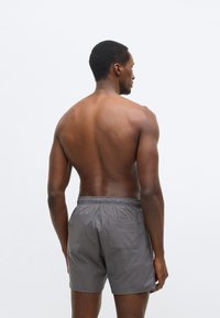 Sive kupaće gaće s elastičnim pojasom i bočnim džepovima, izrađene od laganog materijala, prikazane s boka na neutralnoj pozadini.