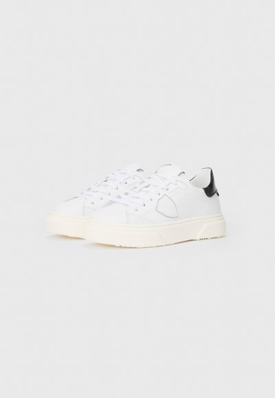 Weiße Sneakers mit glatter Textur, ausgestattet mit einem schwarzen Akzent an der Ferse, flachen weißen Schnürsenkeln und einer massiven weißen Sohle.