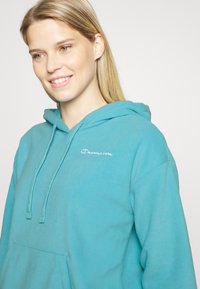 Hoodie azul claro feito de um tecido suave, apresentando um bolso frontal e um logótipo bordado em branco. Inclui cordões ajustáveis na capuz.