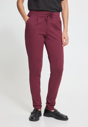 Femme portant un pantalon slim bordeaux avec taille élastiquée à cordon, des mocassins noirs et une chemise noire, debout avec une main dans la poche.