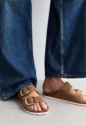 Brown sandals with two adjustable buckles, perforated upper design, cork footbed, and white rubber sole. Paired with loose denim jeans.