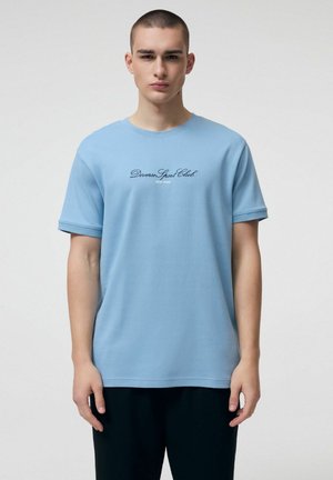 Młody mężczyzna z krótkimi włosami, ubrany w jasnoniebieską koszulkę z napisem "Diverse Sport Club New York", stoi na tle jasnego, jednolitego tła.