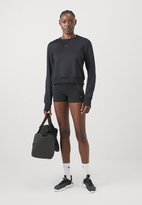 Zwarte Nike-sweater met een ronde halslijn, gecombineerd met zwarte shorts met zijpanelen van gaas. Model houdt een zwarte sporttas vast, draagt witte sokken en zwarte schoenen.