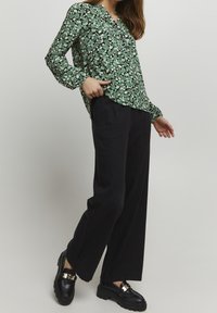 Blusa floreale verde con maniche lunghe e scollo a V, abbinata a pantaloni neri a gamba larga e mocassini neri spessi con accenti dorati.