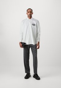 Chemise blanche à boutons avec des manches longues et un logo noir. Pantalon gris à motif et baskets noires. Tient un petit sac noir.