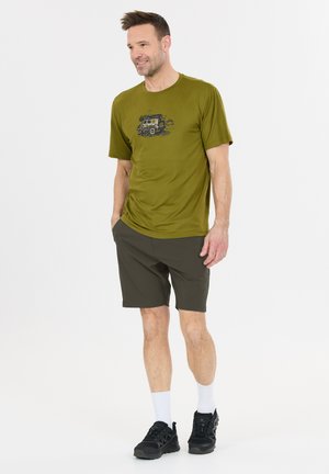 Man draagt een olijfgroen T-shirt met een voertuiggrafiek, donkere korte broek, witte sokken en zwarte sneakers, staand met handen in de zakken.