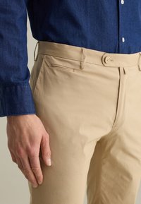 Beige broek met een gladde textuur, voorzien van een frontzak en een knoopsluiting. Een hand rust voorzichtig op de stof, wat de details toont.