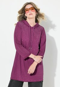 Violetti hupparitunika pitkällä hihalla, jossa on koristeellisia niittejä yläosassa ja kiristysnauha. Pehmeä rakenne ja rento malli.