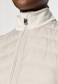 Lys beige quiltet jakke med høy krage, glidelås-lukking, og metallisk glidelåskroker med et fremtredende bokstav B. Jevn tekstur.