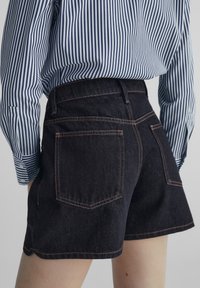Mörka denimshorts med hög midja, med kontrasterande orange sömmar och två bakfickor, tillsammans med en blårandig skjorta.