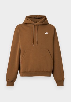 Młody mężczyzna z fryzurą cornrows, ubrany w brązową bluzę z kapturem Nike oraz jasnoniebieskie, wyblakłe jeansy, stoi na jednolitym białym tle.