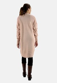 Lång, ljusrosa stickad tröja med texturerade, bubbliga ärmar, ribbade manschetter och en avslappnad silhuett. Buren över svarta leggings och bruna stövlar.