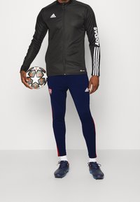 Černá sportovní bunda s bílým logem Adidas, se zipem vpředu a třemi bílými pruhy na rukávu. Námořnické kalhoty s červenými akcenty. Drží fotbal.