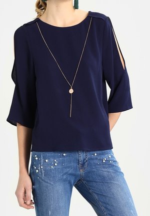 Femme portant un chemisier bleu marine avec des découpes aux épaules et des manches mi-longues, associé à un jean bleu orné de perles éparses.