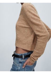 Hellbrauner Strickpullover mit langen Ärmeln, kurz geschnittenem Design und subtiler Textur, kombiniert mit hoch taillierten blauen Jeans und einem schwarzen Gürtel.