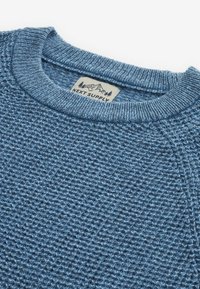 Gebreide trui in lichtblauw met een textuurpatroon. Heeft een ronde hals en een label met zichtbare tekst "NEXT SUPPLY". Zachte garenafwerking.