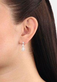 Orecchino a goccia d'argento con cristalli a goccia e rotondi indossato all'orecchio sinistro di una donna con capelli castano scuro raccolti.