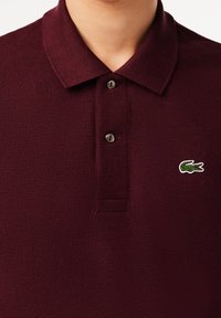 Polo bourgogne avec un tissage texturé, deux boutons au col, et un logo de crocodile vert sur la poitrine gauche.