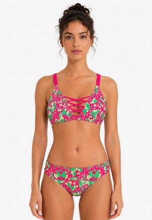 Woman with curly hair tied up wearing a pink, green, and beige patterned bikini with crisscross straps on top, standing against a plain background.