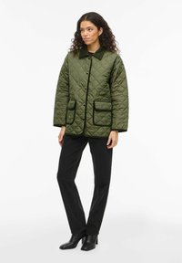 Veste matelassée vert olive avec col, bordure noire et deux poches avant, associée à un pantalon noir et des bottes à talons.