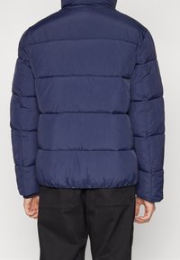 Marinblå vadderad jacka med ett kviltat mönster, som har horisontella sömmar och en hög krage. Tillverkad av lätt, vattentåligt material.