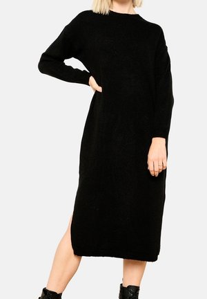 Vestido de punto negro con mangas largas, corte relajado, aberturas laterales, dobladillo acanalado y una textura suave, alcanzando una longitud hasta la mitad de la pantorrilla.