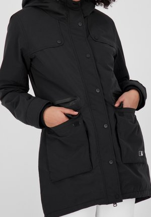 Manteau d'hiver noir à capuche avec boutons-pression, deux grandes poches à l'avant, mains glissées à l'intérieur des poches, porté par-dessus un pantalon blanc.