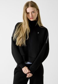 Czarna, krótka bluza na zamek z długimi rękawami, z małym logo na piersi i kontrastującymi białymi paskami na ramionach.