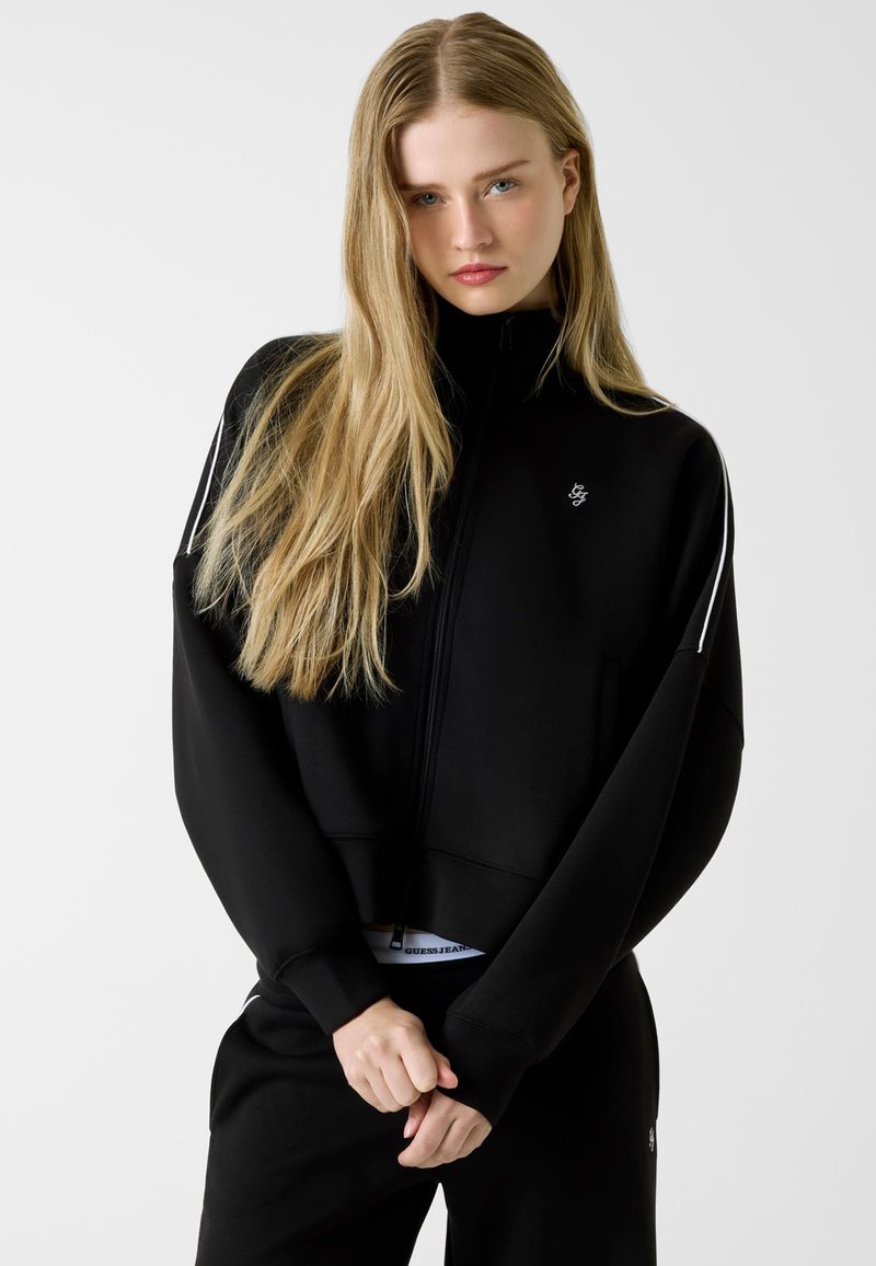 Czarna, krótka bluza na zamek z długimi rękawami, z małym logo na piersi i kontrastującymi białymi paskami na ramionach.
