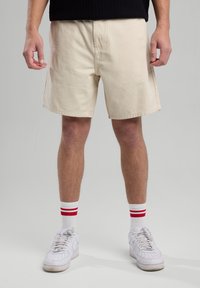 Pantaloni corti in cotone beige con una vestibilità rilassata, davanti piatto, tasche laterali e orli rifiniti, abbinati a sneakers bianche e calze a righe rosse.