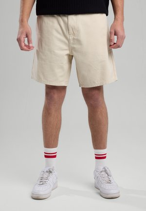 Beige cotton shorts with a relaxed fit, flat front, side pockets, and finished hems, paired with white sneakers and red-striped socks.