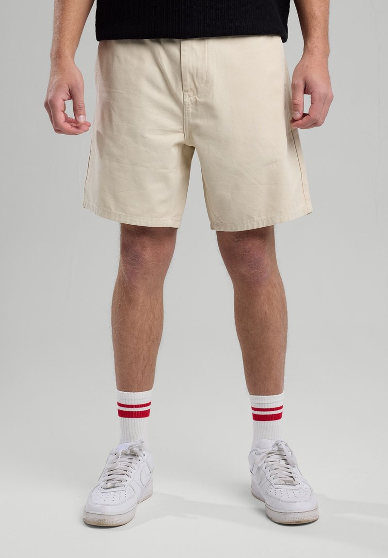 Pantaloni corti in cotone beige con una vestibilità rilassata, davanti piatto, tasche laterali e orli rifiniti, abbinati a sneakers bianche e calze a righe rosse.