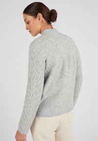 Suéter de punto gris claro con patrón de trenzas en los hombros y las mangas, textura acanalada y cuello alto. Combinado con pantalones color crema.