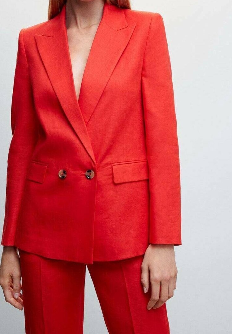 Donna che indossa un vivace blazer rosso a doppio petto e pantaloni abbinati, in piedi di fronte a uno sfondo neutro.