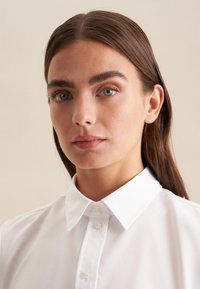 Camicia bianca con colletto classico, caratterizzata da una texture liscia e un design aderente, che presenta un'estetica pulita e semplice.