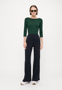 Haut à manches longues vert foncé, décolleté rond, associé à un pantalon large à côtes noir. Le modèle porte des lunettes de soleil rectangulaires et des chaussures blanches.