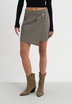 Mujer con falda envolvente de cuadros marrones con hebilla, camiseta de manga larga negra acanalada y botas de vaquero de ante color marrón claro, de pie sobre un suelo blanco.