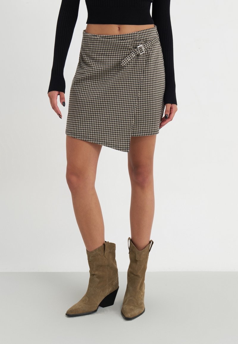Femme portant une jupe portefeuille à carreaux marron avec boucle, un haut à manches longues côtelé noir et des bottes de cowboy en suède beige, debout sur un sol blanc.