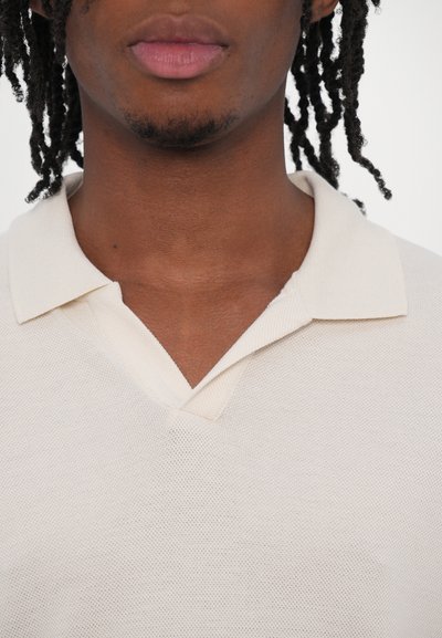Polo shirt en maille gaufrée beige clair avec un col en V et un col court. Le tissu a une apparence texturée et une finition douce.