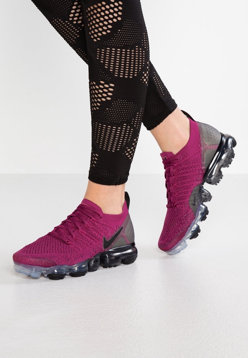 Jambes en leggings noirs à motifs portant des baskets Nike violettes avec des semelles transparentes à bulles sur un sol blanc.